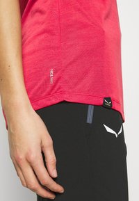 Camisa roja de manga corta hecha de un tejido ligero que presenta una etiqueta "DRYTON" en el lateral; combinada con pantalones negros que muestran un logo blanco y un acento.