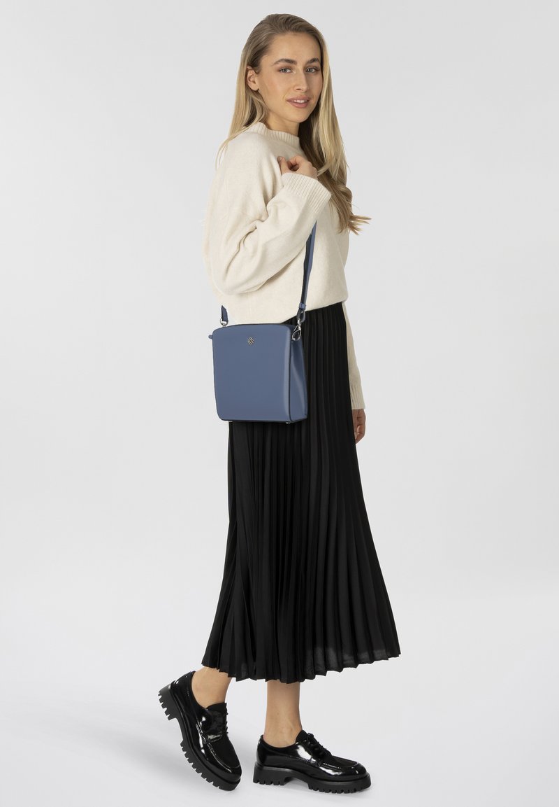 Lichtblauwe synthetische crossbodytas met een vierkante vorm, afneembare band en een gouden logo aan de voorkant. Draagt bij een crèmekleurige trui en een zwarte, plissé rok.