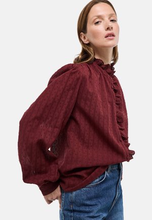 Blouse bordeaux en tissu texturé, avec un col à volants, des rayures verticales et des manches bouffantes. Portée avec un jean en denim bleu.