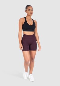 Zwarte sportbeha en bordeauxrode korte broek, beide gemaakt van rekbare stof. Model staat op een neutrale achtergrond en draagt witte sneakers.