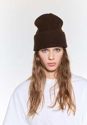 Bruine geribbelde gebreide beanie zit op het hoofd, met een omgeslagen rand. Het model draagt een loszittend wit shirt met lang, golvend haar dat het gezicht omlijst.