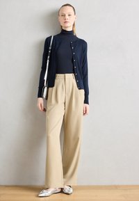 Cardigan marine avec boutons dorés, assorti à un col roulé noir ajusté et un pantalon beige à jambes larges. Des ballerines argentées complètent le look.