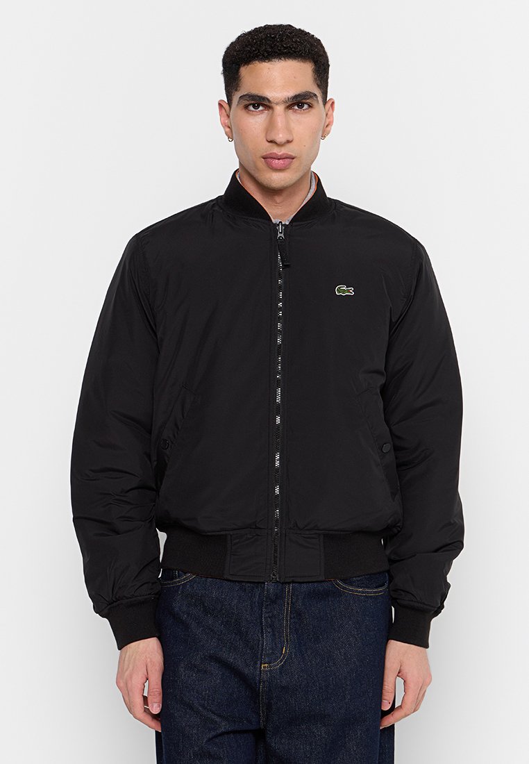 Veste bomber noire en nylon lisse, avec fermeture éclair, poignets et ourlet côtelés, et un petit logo vert sur la poitrine gauche.