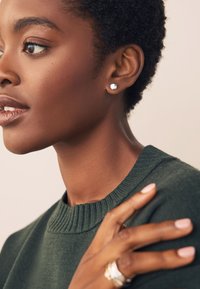 Dark green, ribbed sweater; close-up of a face with short, curly hair; silver earrings; hand with multiple rings resting on the shoulder.