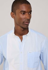 Camicia button-up blu chiaro e bianca con righe verticali, taschino sul petto con logo e un design del colletto classico.