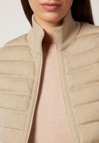 Nainen, jolla on päällään vaalean beige vuorattu takki, jossa on ribbattua kangasta oleva korkea kaulus, sekä vaaleanpunainen neule.