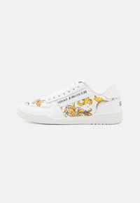 Weiße Sneaker mit aufwendig gestaltetem gelb-goldenem Barockmuster, beschriftet mit "Versace Jeans Couture." Verfügt über ein glattes Obermaterial aus Leder und eine Gummisohle.