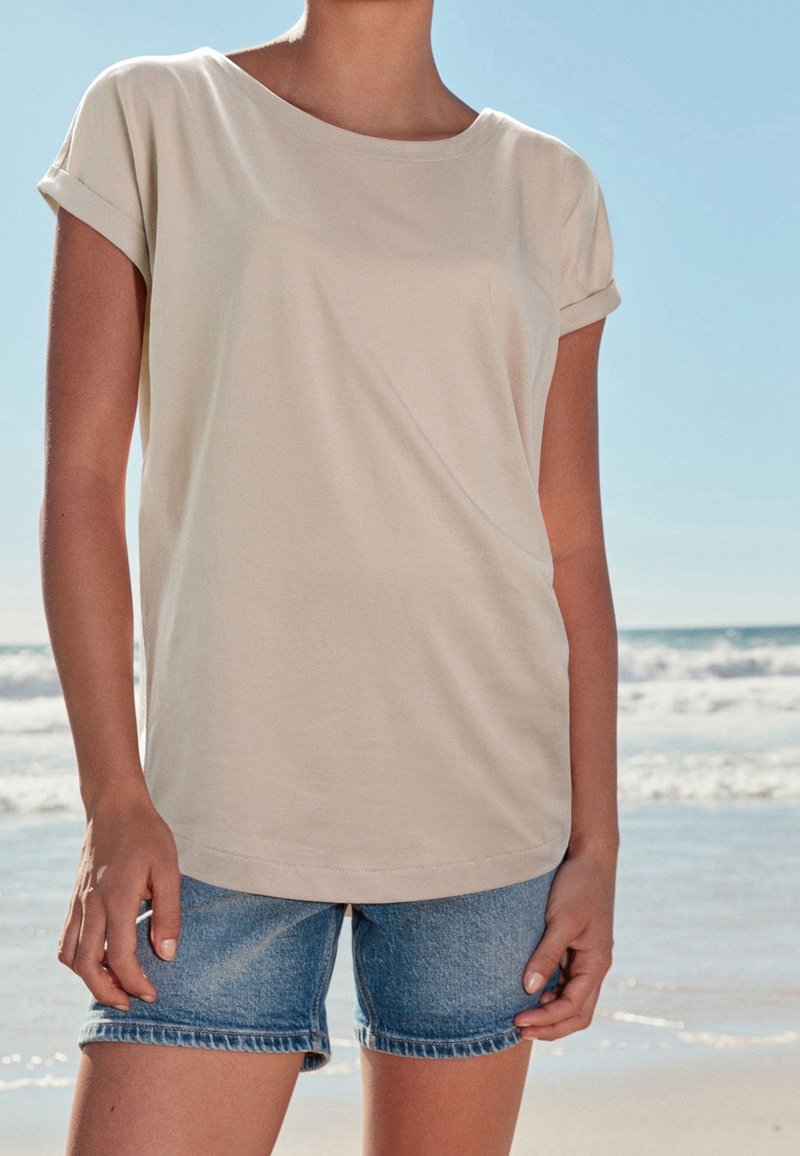 Personne portant une chemise beige à manches courtes et un short en jean bleu, debout sur une plage ensoleillée près de l'océan.