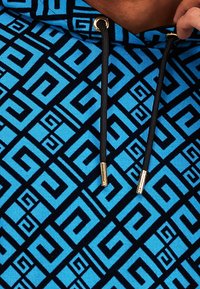 Modrá mikina s kapucí a černým geometrickým vzorem. Má černé šňůrky s zlatými koncovkami. Měkká texturovaná látka.