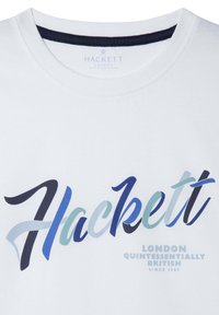Camiseta de algodón blanca con un cuello circular en color navy. En la parte frontal se muestra un logo "Hackett" en un degradado de tonos azules con texto adicional debajo.
