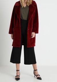 Vrouw in een dieprode lange bontjas, olijfgroen blouse, zwarte wijdvallende cropped broek en zwarte enkellaarsjes met enkelband op een witte achtergrond.