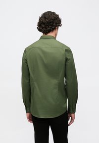 Dlouhý rukáv zelené košile z hladké látky. Má klasický límec, manžety na knoflíky a přizpůsobený střih, viditelný ze zadní strany.