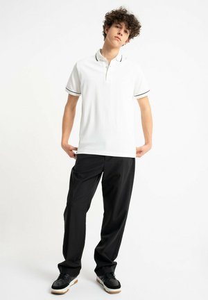 Młody mężczyzna ubrany w biały polo z czarnym wykończeniem, czarne spodnie i czarne trampki, stojący na jednolitym białym tle.