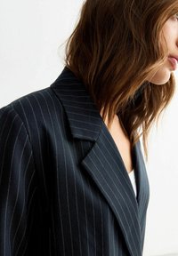 Blazer blu navy con righe sottili, colletto a rever, texture morbida e vestibilità su misura, mostrato di lato con una maglietta bianca sotto.