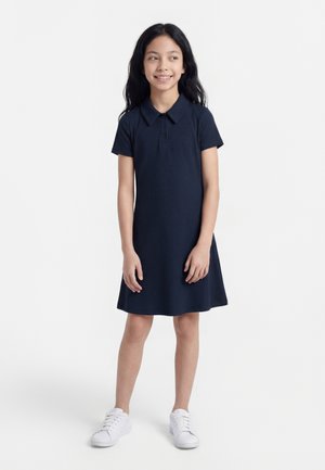 Junges Mädchen mit langem dunklem Haar, das ein marineblaues kurzärmeliges Kleid und weiße Turnschuhe trägt, steht und lächelt leicht, während sie nach rechts schaut.