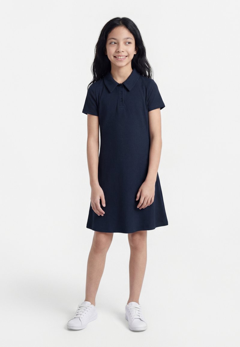 Νεαρή κοπέλα με μακριά σκούρα μαλλιά, φορώντας ένα μπλε σκούρο φόρεμα με κοντά μανίκια και λευκά αθλητικά παπούτσια, όρθια και χαμογελώντας ελαφρώς, κοιτάζοντας προς τα δεξιά.