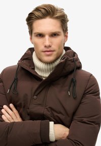 Chaqueta de plumón marrón con capucha ajustable, con cierres de cremallera y botón. Superpuesta con un suéter de cuello alto de punto ribeteado en color crema.