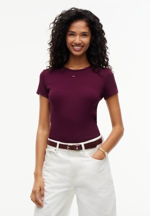 Femme souriante aux cheveux bouclés portant un haut moulant violet foncé à manches courtes, un pantalon blanc, une ceinture marron et une montre en or, debout avec une main dans la poche.