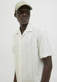 Chemise à manches courtes, avec col en tissu léger blanc, ornée de rayures verticales et de subtils accents texturés. Une casquette vert clair complète le look.
