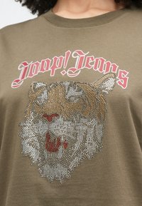 Artimos nuotraukos alyvuogių spalvos marškinėliai su tigrinio veido, puošto cirkoniu, atvaizdu ir viršuje rožinėmis raidėmis „Joop! Jeans“.