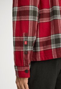 Czerwona i czarna koszula flanelowa w kratę z guzikami na mankietach oraz czerwoną etykietą akcentującą z wykrzyknikiem. Miękka tekstura i swobodny design.