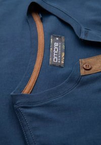 Camisa de color azul marino con detalles en marrón en el cuello y la zona de los botones; presenta una etiqueta con el texto "Ombre Quintessence". Textura de tela suave.