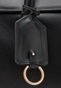 Bolso de piel negra con acabado texturizado, que presenta una etiqueta rectangular y un anillo de metal de color dorado como adorno.