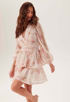 Kobieta z długimi brązowymi włosami, ubrana w jasnoróżową, kwiatową sukienkę kopertową z bufkami na długich rękawach i warstwową spódnicą, pozująca na jednolitym tle.