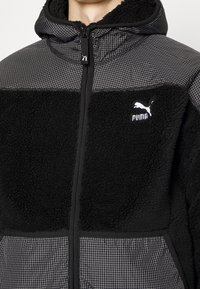 Sort fleecejakke med rute-mønstrede stofpaneler, fuld lynlås og hvid Puma-logo på venstre bryst.