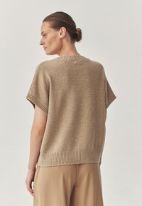Maglione beige a maniche corte lavorato a maglia con un motivo testurizzato, collo e orlo a coste, indossato con pantaloni di colore chiaro.