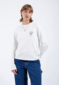 White long-sleeve shirt featuring a black heart and peace symbol design on the front. Paired with blue denim jeans.