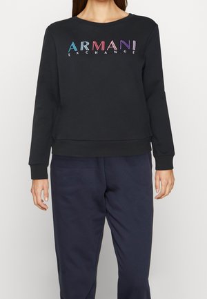 Kvinde iført en sort langærmet sweatshirt med flerfarvet "ARMANI EXCHANGE" tekst, kombineret med mørkeblå bukser.