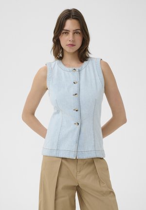 Femeie în picioare cu mâinile la spate, purtând un top fără mâneci din denim deschis la culoare cu nasturi și pantaloni bej pe un fundal simplu.