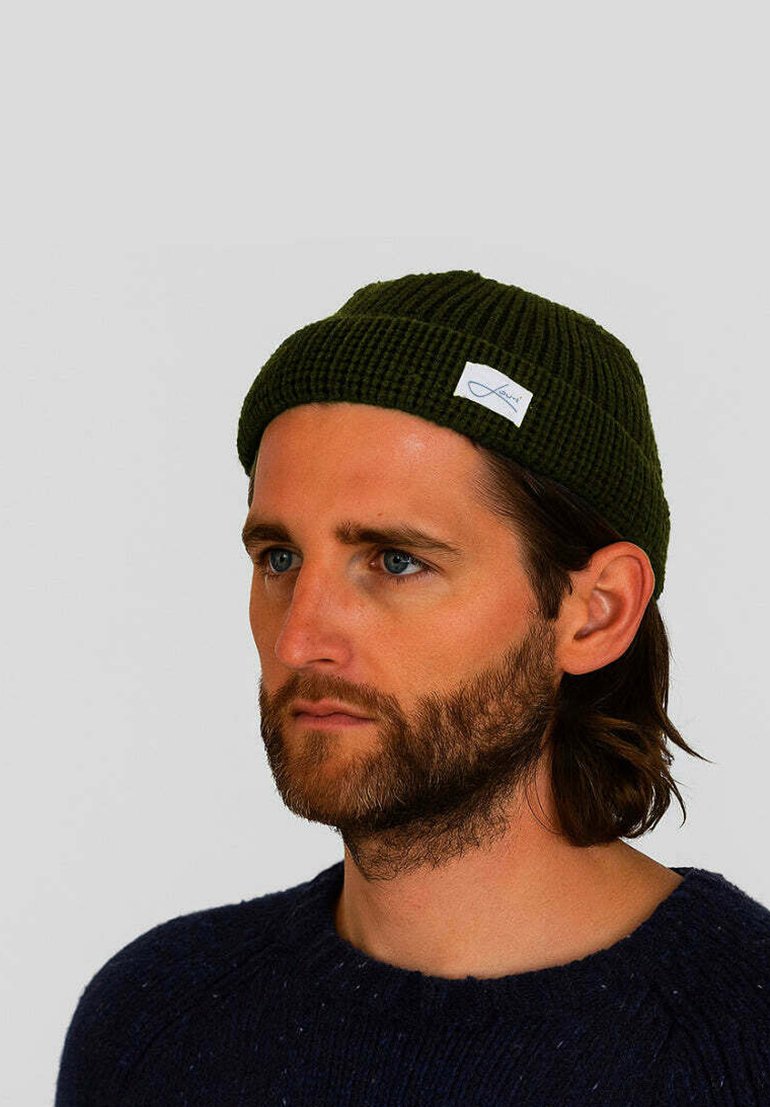 Dark green knitted beanie hat with a ribbed texture and a small white logo tag. Worn on a person with light clothing and a beard.