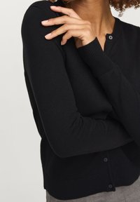 Svart stickad kofta med långa ärmar, ribbade ärmslut och rund halsringning. Har knäppning med knappar och en mjuk textur.