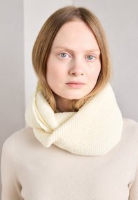 Bílý pletený šálu s strukturou a vzorem herringbone, nošený volně kolem krku přes béžový svetr. Měkký a útulný vzhled.