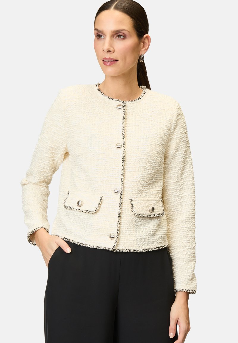 Cremefarbene Tweedjacke aus strukturiertem Stoff, runder Ausschnitt, silberne Knöpfe und zwei Fronttaschen. Mit schwarzen Akzenten verziert.