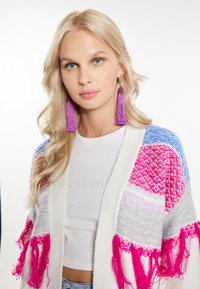 Stickad cardigan med färgglada mönster i rosa, grått och blått; har fransar. Modellen bär en vit topp och lila tofsörhängen.