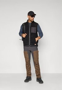 Fekete polármellény szürke zsebbel, kék ing fölött viselve. Barna cargo nadrág térdvédőkkel. Fekete sapka és napszemüveg, hátizsákkal.