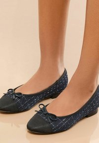 Zwei schwarze und marineblaue Ballerinas mit einem strukturierten Design und einer Schleifendetail, präsentiert an Beinen mit einem glatten Hautton vor einem neutralen Hintergrund.