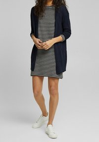 Marineblauwe lange cardigan over een gestreepte zwart-witte jurk. De jurk heeft korte mouwen, een ronde halslijn en is knielang. Witte sneakers.