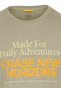 Lys grå t-shirt med korte ærmer og rund halsudskæring. Har gul og orange tekst: "Skabt til Daglige Eventyr" og "FORFOLG NYE HORISONTER."