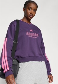 Sudadera morada con rayas rosas en las mangas, con un logo de Adidas bordado. Combinada con pantalones de chándal grises y una bolsa de hombro negra.