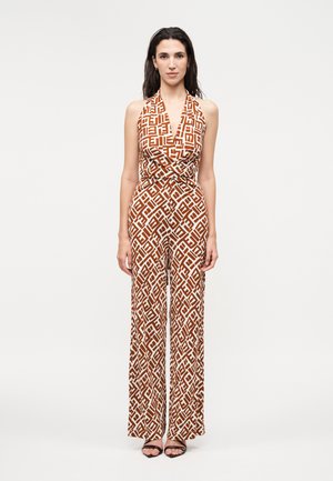 Žena oblečená v hnedo-bielom geometrickom overal s širokými nohavicami a čiernymi otvorenými lodičkami, stojaca pred jednoduchým pozadím.