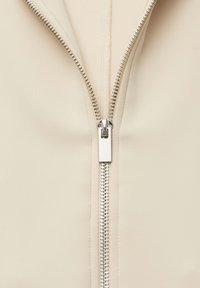 Beige kledingstuk met ritssluiting, voorzien van een metalen rits, gladde stoftextuur en nauwkeurige stiksels langs de randen, gecentreerd op de ritssluiting.