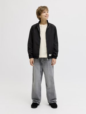 Młoda osoba stoi, ubrana w czarną kurtkę na zamek, białą koszulkę, luźne szare dżinsy i czarne buty, patrzy lekko na bok z uśmiechem.