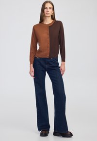 Bruine en donkerbruine button-up cardigan met ronde hals, ribbelstructuur, gedragen met donkerblauwe flared jeans en chunky zwarte schoenen.