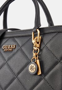 Sac à main noir matelassé avec ferrures dorées, comprenant un pendentif avec logo et une petite étiquette. Fabriqué en matériau synthétique texturé.