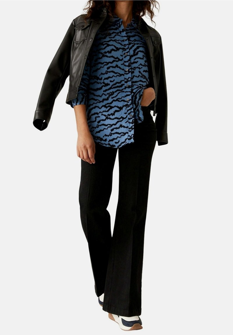Femme portant une chemise à motifs bleus à boutons, une veste en cuir noire posée sur les épaules, un pantalon noir et des baskets blanches avec des accents bleu marine.