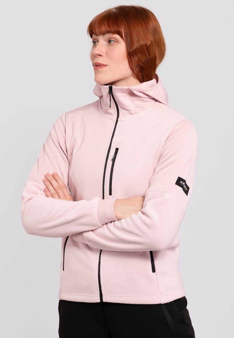 Veste en polaire rose clair avec un col haut, fermeture éclair frontale et accents de fermeture éclair noirs. Logo notable sur la manche gauche.